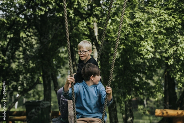 Fototapeta Two boys sit on a rustic wooden swing suspended by ropes in a lush green park surrounded by trees and wooden benches, enjoying a peaceful moment outdoors.