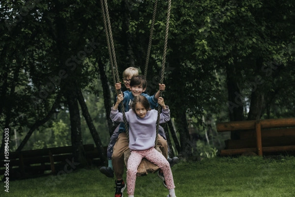 Fototapeta Three children sit close together on a wooden rope swing in a wooded area, laughing and playing near a carved wooden bear statue. An energetic and playful scene. 