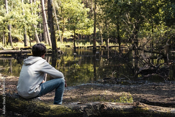 Fototapeta A boy rests on a tree trunk, gazing at the tranquil pond in the forest. The warm sunlight filters through the trees, creating a serene atmosphere filled with nature