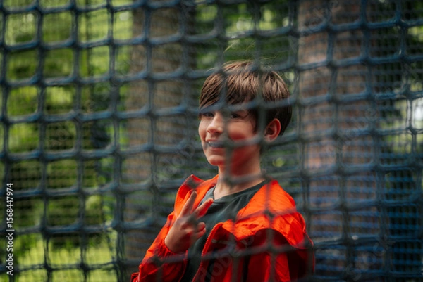 Fototapeta A cheerful boy in an orange jacket poses with a peace sign behind a blue safety net outdoors, in a playful atmosphere, surrounded by greenery.
