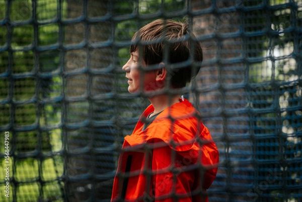 Fototapeta A young boy in a bright orange jacket smiles in profile behind blue netting outdoors, with trees and soft natural light in the background.