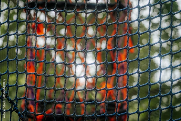 Fototapeta A detailed view of a blue net with a soft-focus background showing blurred orange lights or fabric and green bokeh from trees.