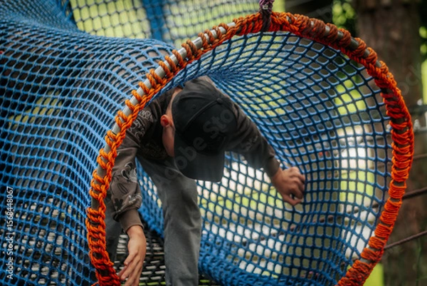 Fototapeta A little boy in a hat and hoodie climbs through a bright blue and orange rope tunnel at an outdoor playground.