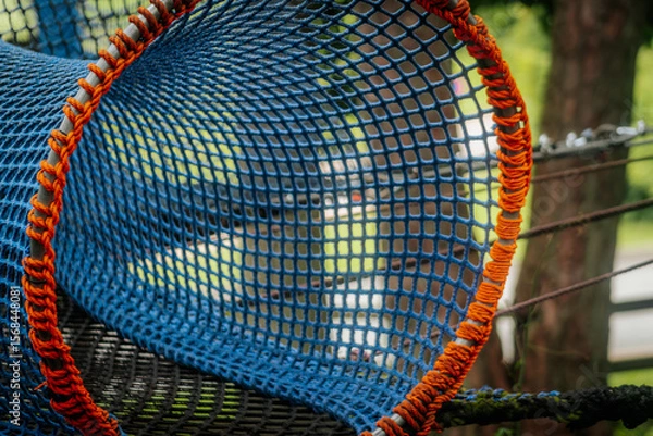 Fototapeta An empty blue-orange rope tunnel suspended in a wooded outdoor playground, forming a curved, mesh structure.