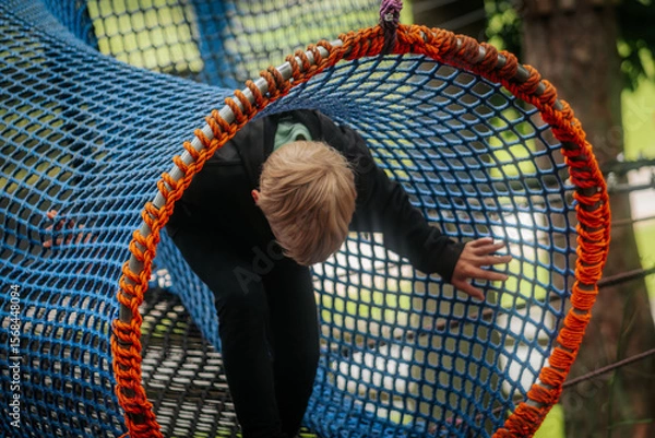 Fototapeta A small child in a black hoodie moves through a colorful rope tunnel made of blue and orange netting in a forest adventure park.