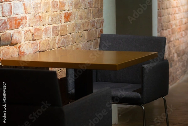 Fototapeta A quiet, stylish lounge area in a cafe with a mustard-colored table, dark upholstered chairs, and exposed brick walls in soft lighting.
