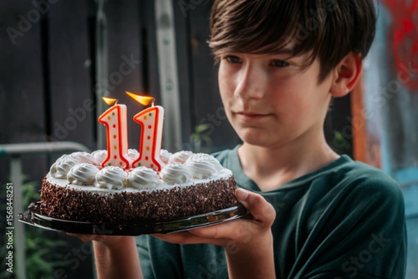 Fototapeta A boy holds a decorated strawberry cheesecake cake with chocolate on the edge. The 11th candle is lit on top of the cake, celebrating his 11th birthday. The background is softly blurred, highlighting 