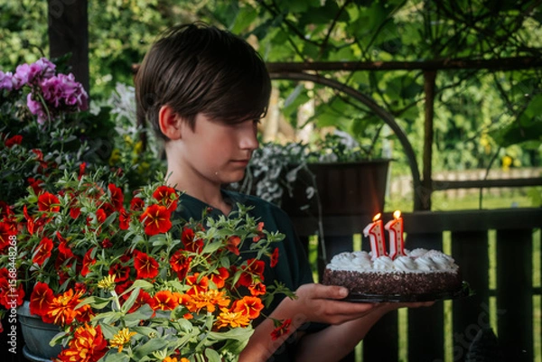 Fototapeta A boy holds a decorated strawberry cheesecake cake with chocolate on the edge. The 11th candle is lit on top of the cake, celebrating his 11th birthday. The background is softly blurred, highlighting 