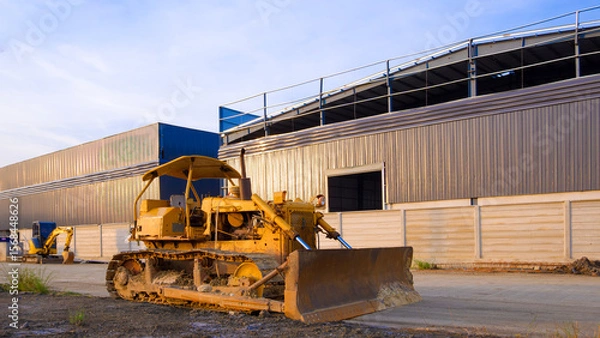 Fototapeta The old bulldozer tractor on the ground in front of industrial warehouse factory building structure in construction site, perspective side view