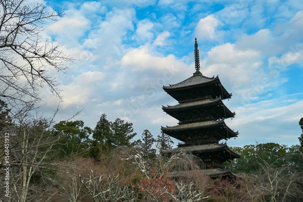 Fototapeta Magnificent five-story wooden pagoda of Ninna-ji Temple towers above the surrounding bare winter trees. Ancient, intricate structure set against a dynamic blue sky with fluffy white clouds in Kyoto.