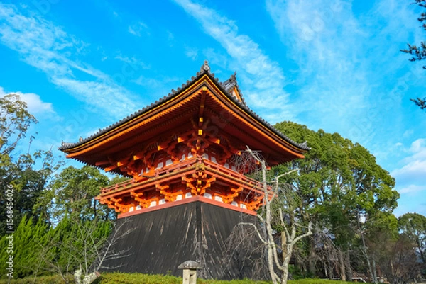 Fototapeta The brightly painted orange and white Kyozo, or sutra repository, at Ninna-ji Temple in Kyoto stands out with its unique architectural style against a backdrop of green trees and a clear blue sky.