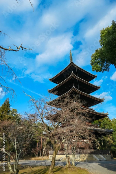 Fototapeta Impressive five-story wooden pagoda of Ninna-ji Temple in Kyoto stands tall against a vivid blue sky with scattered white clouds, its intricate design framed by bare winter branches, seen from below.