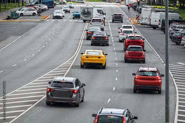 Fototapeta Urban traffic scene on a multi-lane road during the day. Various cars drive along a wide boulevard surrounded by parked cars. Road markings and a red traffic light are visible in the distance