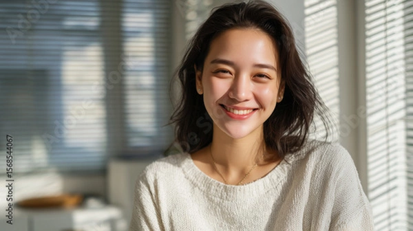 Obraz Indoor portrait of cheerful Asian woman with radiant smile and straight black hair cascading over her shoulders, sunlight streaming through window blinds, minimalistic workspace be