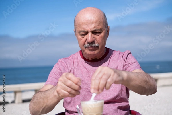 Fototapeta Elderly Man Adding a Lot of Sugar to Coffee While Sitting on a Summer Terrace