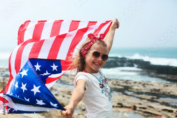 Obraz American flag. Little smiling  patriotic girl with long blond hair, red head band  bandana and sunglasses holding an american flag waving in the wind on the ocean beach. National 4 july. Memorial day