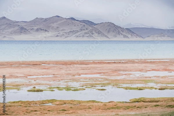 Fototapeta Rocky shore of Lake Karakul with blue water and mountain peaks in the background, cloudy weather and sky with clouds and clouds in the Pamirs