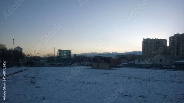 Obraz Snow-covered field and buildings under a cold, early morning sky.