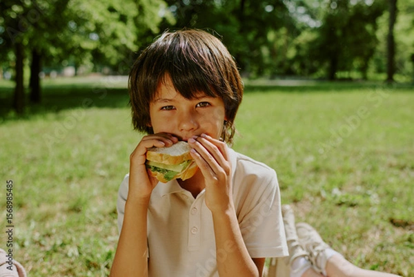 Obraz Portrait of biracial boy child sitting outdoors in park holding sandwich with both hands, eating lunch looking at camera surrounded by green grass and trees in background