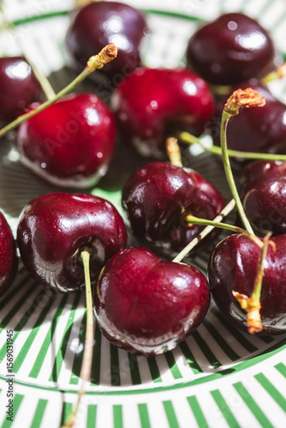 Fototapeta Close-up of fresh dark red cherries with stems on a decorative green striped plate, captured in natural sunlight with vibrant detail and summery freshness.
