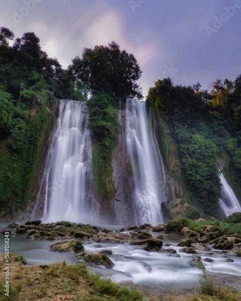 Fototapeta A large, multi-tiered waterfall with a gentle white water cascade, falling from a high cliff. The bottom of the falls features large rocks and water rushing into a clear shallow pool.