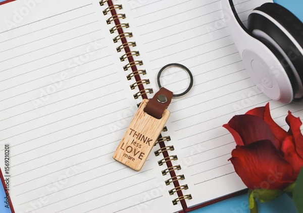 Fototapeta wooden keyring with word "Think less love more" on opened diary  with headphones and red rose  on blue background.