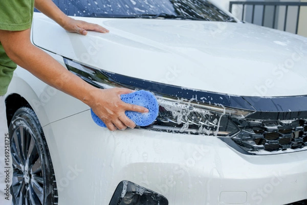 Fototapeta A smiling man is washing his car with soapy water under bright sunlight, showing his enthusiasm and joy in his daily work.