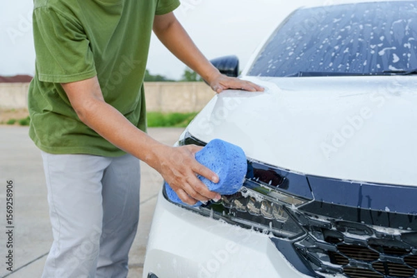 Fototapeta A smiling man is washing his car with soapy water under bright sunlight, showing his enthusiasm and joy in his daily work.