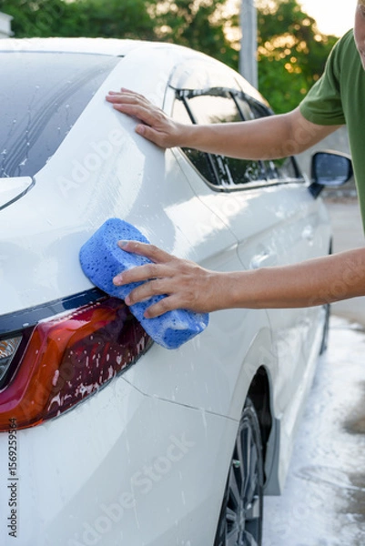 Fototapeta A smiling man is washing his car with soapy water under bright sunlight, showing his enthusiasm and joy in his daily work.