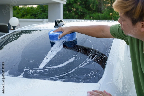 Fototapeta A smiling man is washing his car with soapy water under bright sunlight, showing his enthusiasm and joy in his daily work.