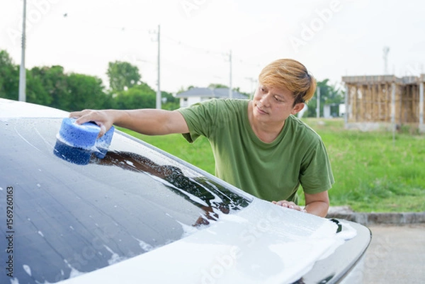 Fototapeta A smiling man is washing his car with soapy water under bright sunlight, showing his enthusiasm and joy in his daily work.