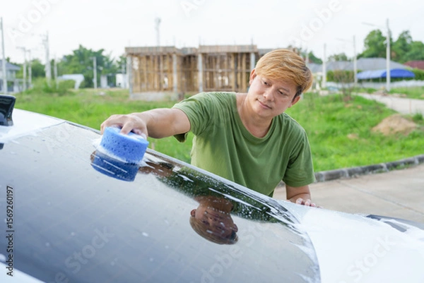 Fototapeta A smiling man is washing his car with soapy water under bright sunlight, showing his enthusiasm and joy in his daily work.
