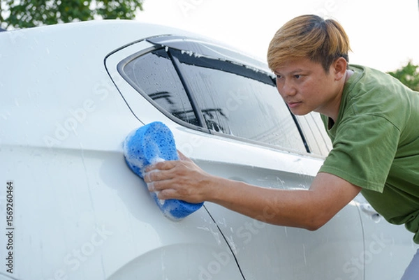 Fototapeta A smiling man is washing his car with soapy water under bright sunlight, showing his enthusiasm and joy in his daily work.
