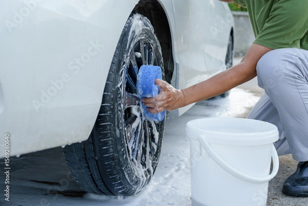 Fototapeta A smiling man is washing his car with soapy water under bright sunlight, showing his enthusiasm and joy in his daily work.
