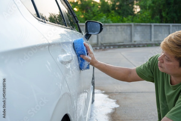 Fototapeta A smiling man is washing his car with soapy water under bright sunlight, showing his enthusiasm and joy in his daily work.