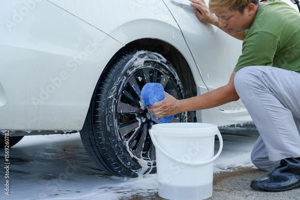 Fototapeta A smiling man is washing his car with soapy water under bright sunlight, showing his enthusiasm and joy in his daily work.