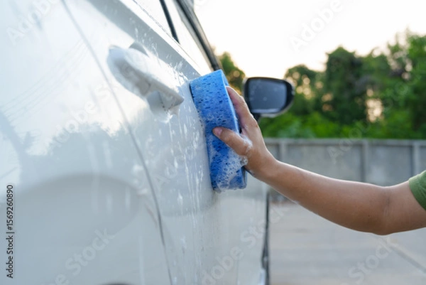 Fototapeta A smiling man is washing his car with soapy water under bright sunlight, showing his enthusiasm and joy in his daily work.