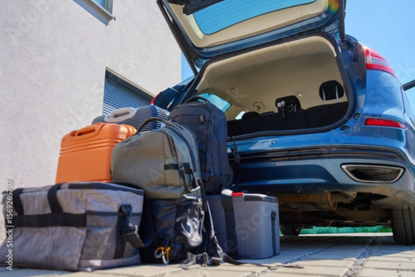 Fototapeta Suitcases and bags in front of open car trunk, standing on pavement near suburban house. Luggage ready for loading in vehicle before travel or moving. Concept of relocation or road trip