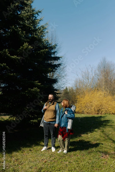 Fototapeta A young couple strolls through the park on a sunny day holding folding chairs in their hands