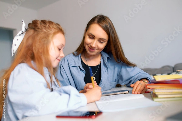 Fototapeta A young schoolgirl and her mother do their homework and write with a pen in a notebook