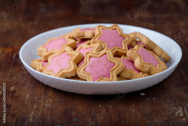 Fototapeta Star shaped cookies with pink frosting on white plate, wooden rustic background. Strawberry icing on cookies in shape of stars. Closeup