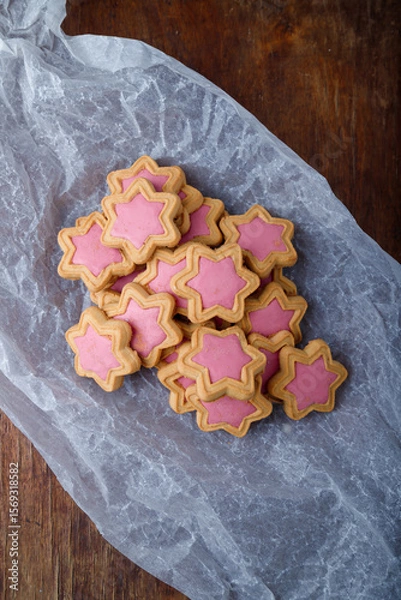 Fototapeta Star shaped cookies with pink frosting on white paper, wooden rustic background. Strawberry icing on cookies in shape of stars. Top view, copy space