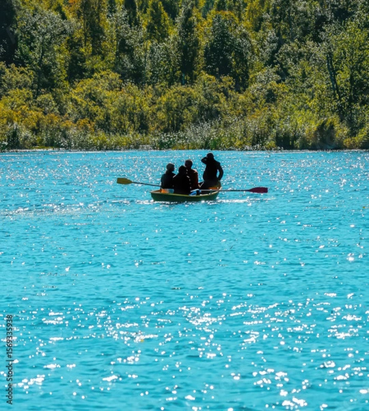 Fototapeta Four people enjoy a sunny day rowing on a vibrant turquoise lake surrounded by lush greenery