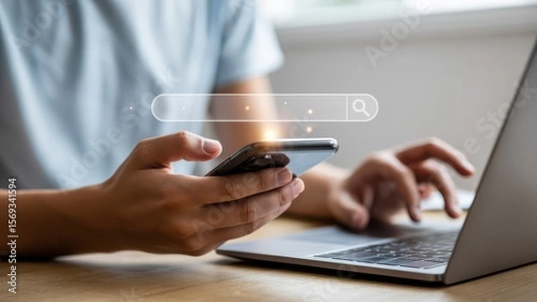 Fototapeta Person using smartphone and laptop with floating search bar icon representing online browsing and digital search technology.