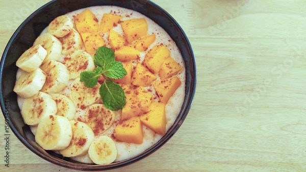 Fototapeta A vegan smoothie bowl with banana and mango slices toppings, sprinkled with cinnamon and garnished with mint, overhead view