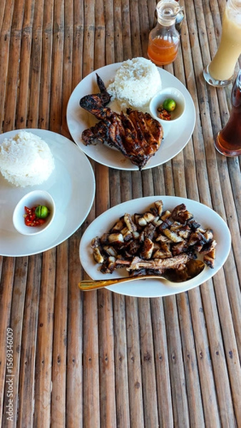 Fototapeta A rustic rural setup with grilled chicken inasal, pork liempo, rice, and iced coffee served in a bamboo table showing an authentic traditional local Filipino food in the Philippines