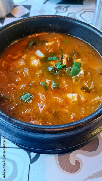Fototapeta A spicy Korean kimchi stew with meat and vegetables served in a black bowl called Kimchi jjigae, creating a warm and savory dining experience