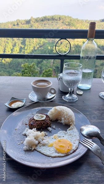 Fototapeta Half-eaten breakfast plate with fried egg, beef patty and rice on a table with an overlooking a lush green landscape, candid moment of a carefree daily life