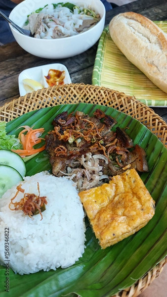 Fototapeta Trung chien or egg omelet, thit heo nuong xa or grilled pork chop and rice on a banana leaf, accompanied by pho bo or beef noodle soup and baguette. Authentic traditional Vietnamese cuisine
