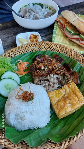 Fototapeta Traditional Vietnamese meal featuring trung chien or omelet, thit heo nuong xa or grilled pork chop and rice on a banana leaf alongside pho bo or beef noodle soup and banh mi or baguette sandwich